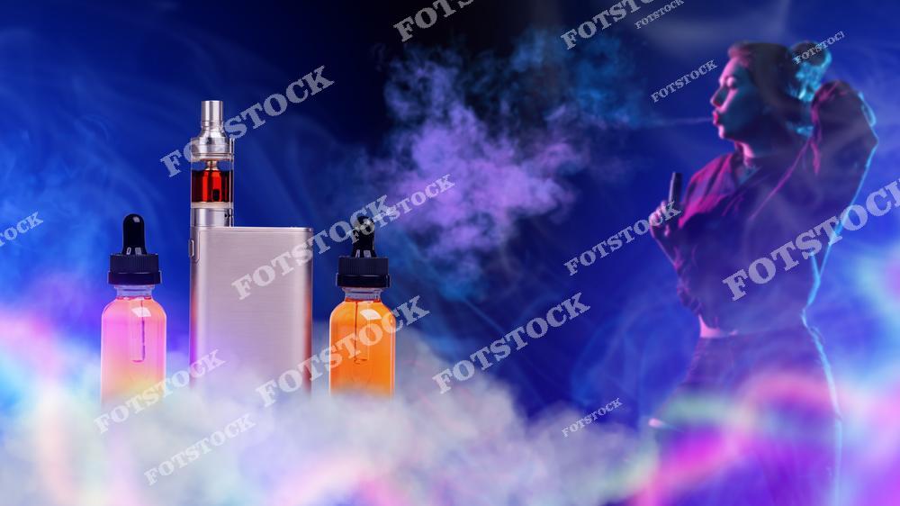Стильное изображение молодой девушки с электронной сигаретой и набором для вейпинга на темном фоне. Женщина-вейпер выпускает густой пар, создавая атмосферную композицию, связанную с современной vape-культурой, lifestyle и альтернативой традиционному курению. Дым, темное освещение и современное устройство для парения подчеркивают концепцию отказа от табака и использования электронных сигарет. Фотография идеально подходит для рекламы vape shop, интернет-магазинов электронных сигарет, жидкостей для вейпа и аксессуаров для парения. Изображение отлично подойдет для маркетинговых материалов, публикаций о вейпинге, молодежной культуре и современных технологиях альтернативного курения.